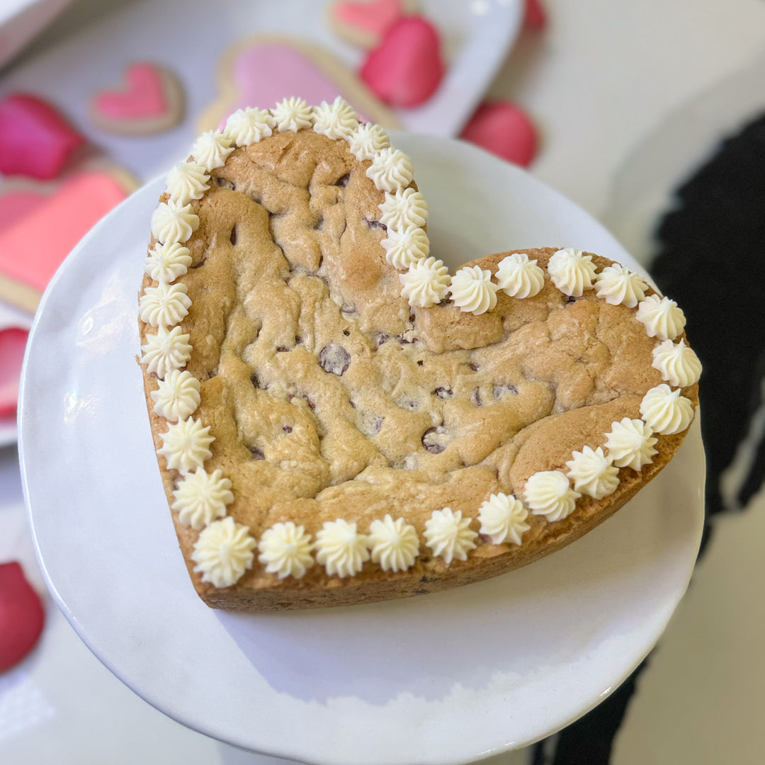 Heart-shaped cookie cake with white frosting on a white plate, surrounded by other cookies and decorations.