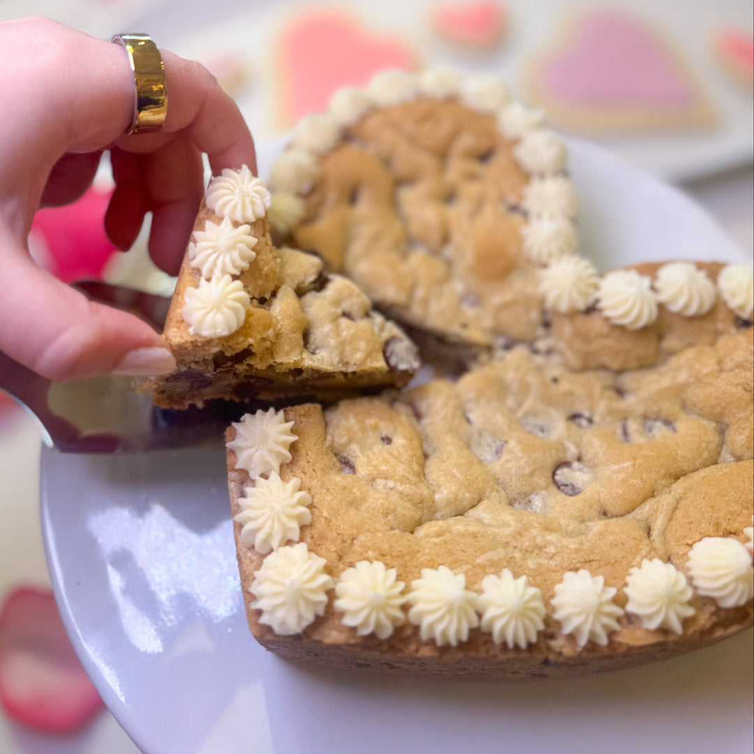 Heart shaped cookie cake with a slice being held by a hand on a decorative tablecloth.